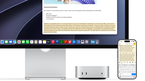Mac mini front, tucked under Studio Display, an iPhone sits to the right of the device and the display, text selected with cut, copy, paste, and AutoFill commands above on iPhone matches highlighted text on display, showcasing Universal Clipboard